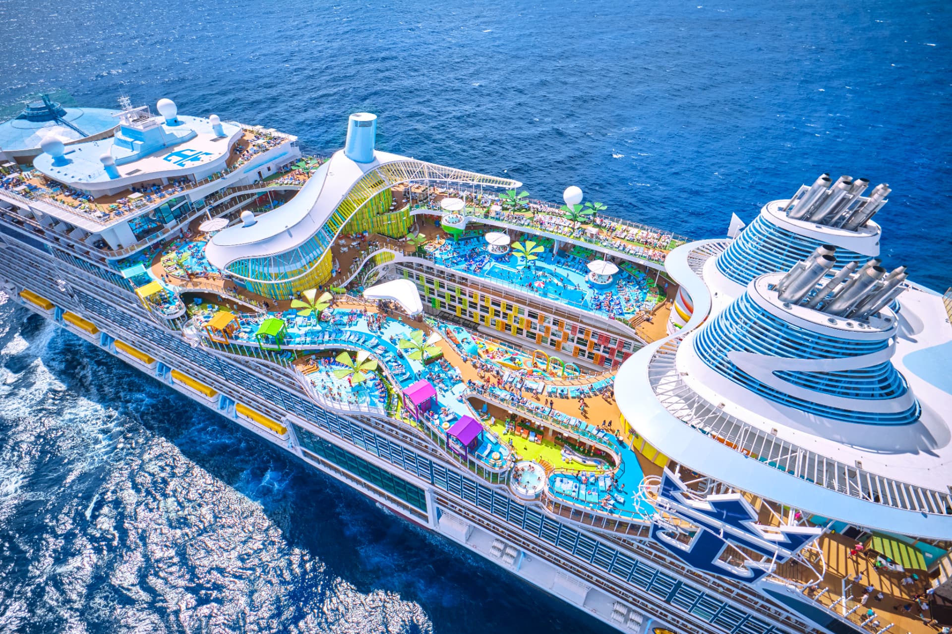 Aerial view of a large Royal Caribbean ship at sea with pools, water slides, and colorful deck areas.