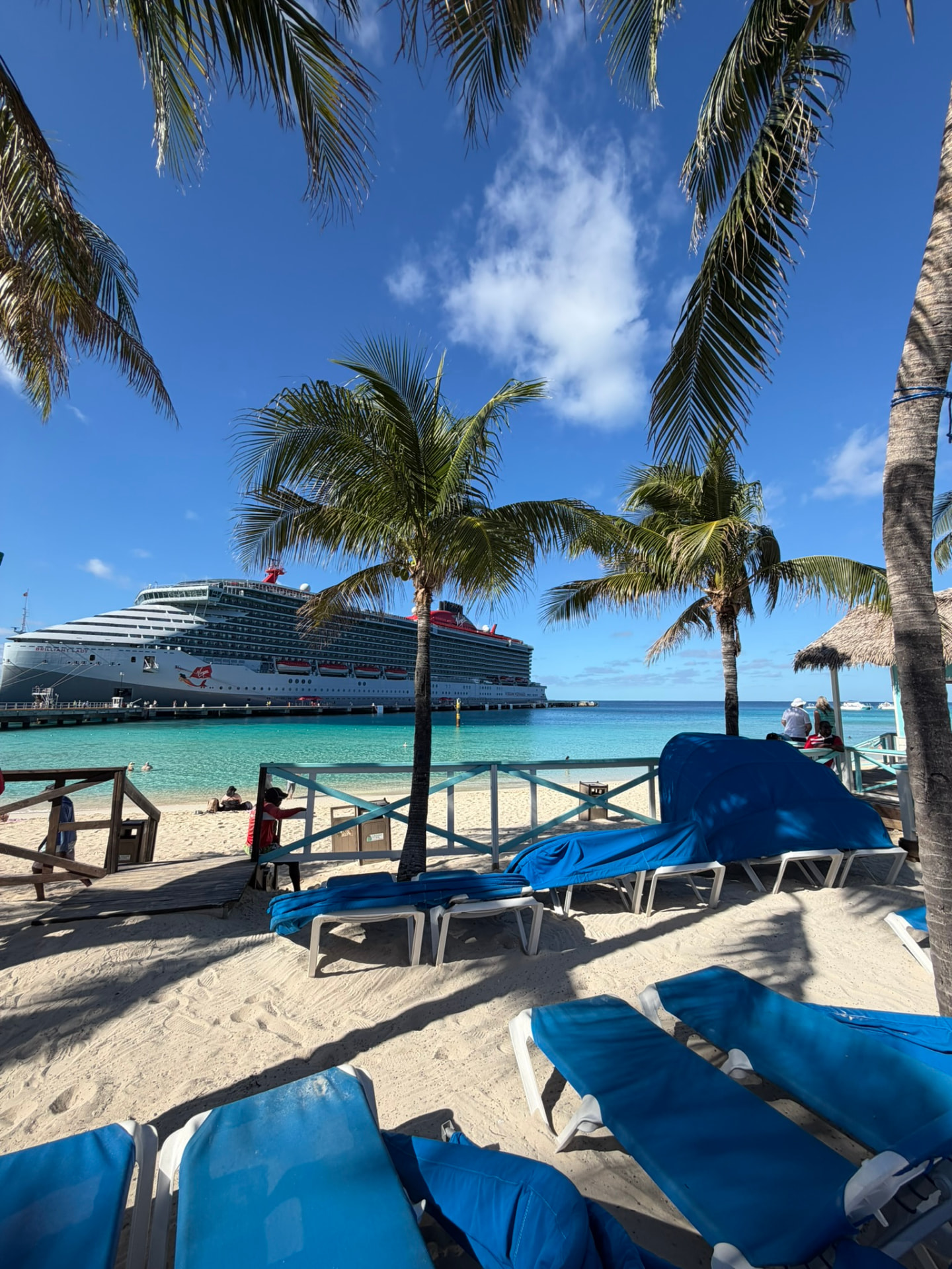 A Virgin Voyages ship docked beyond a palm-lined beach with turquoise water and blue loungers.