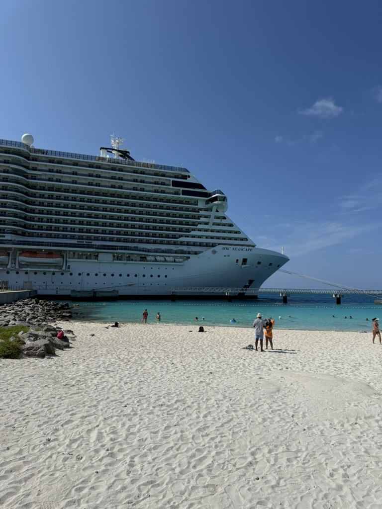 The MSC Seascape docked beside a white-sand beach with turquoise water and swimmers.