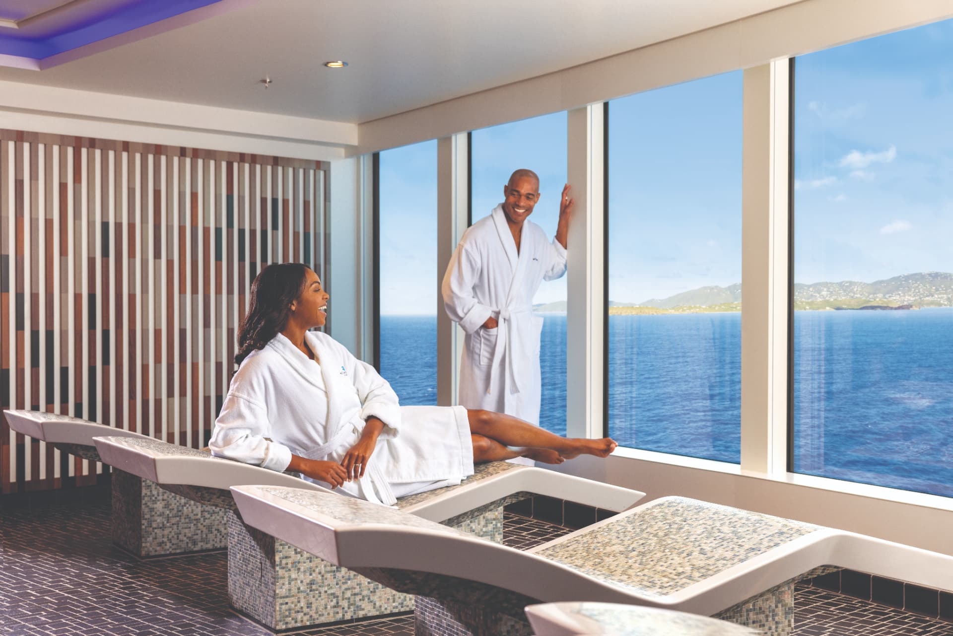 Guests in robes relaxing on heated spa loungers beside floor-to-ceiling ocean windows