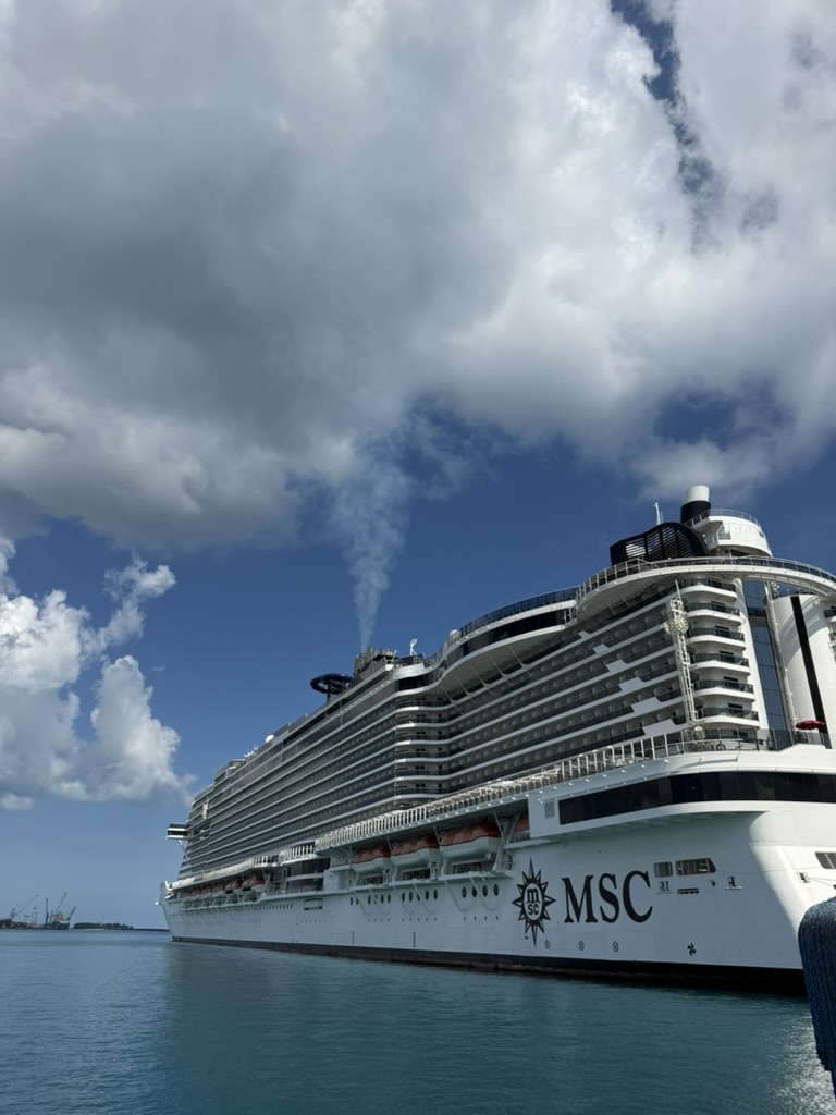 An MSC Cruises ship docked in calm water under tall white clouds.