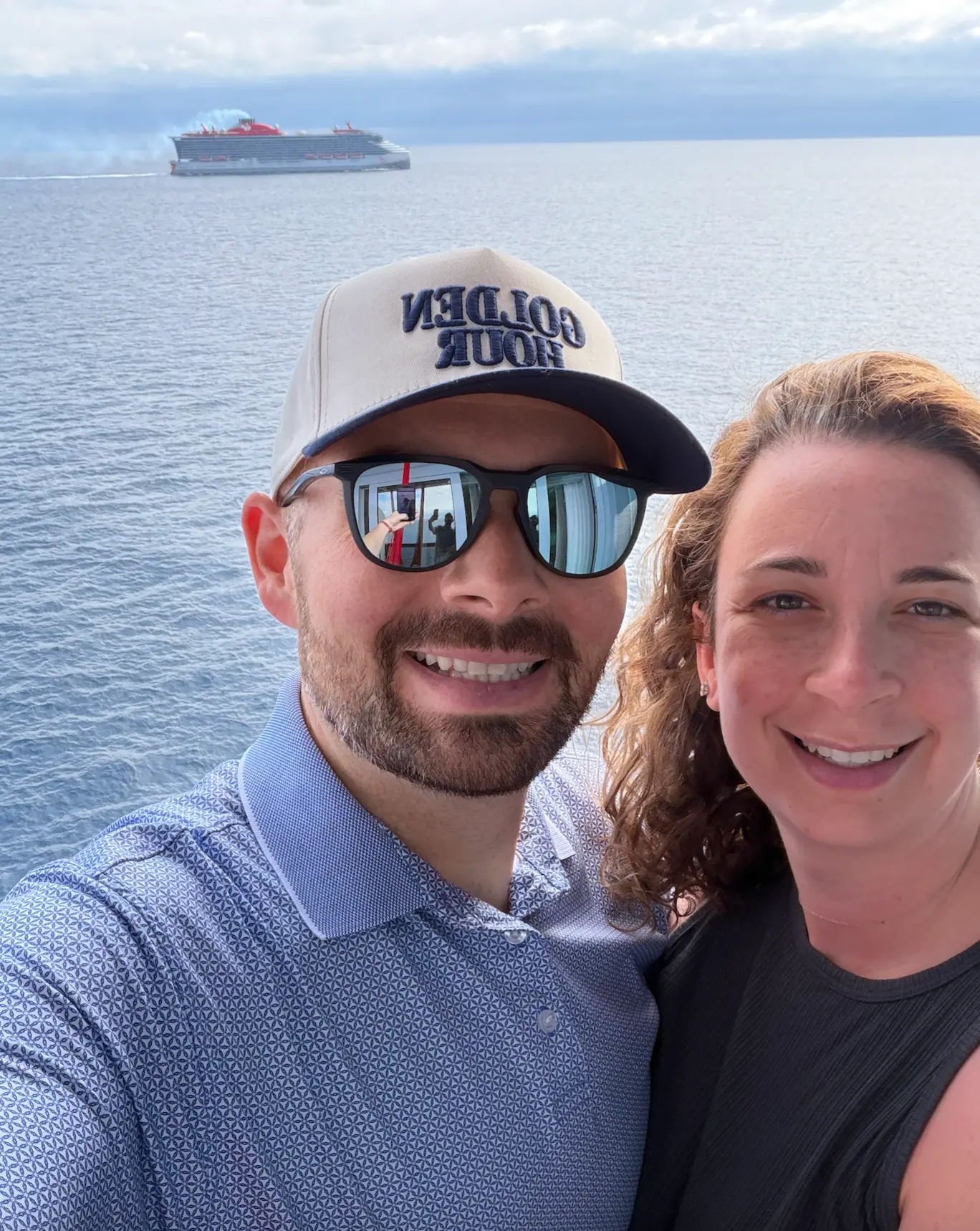 Chris and Emily smiling on a ship balcony with a Virgin Voyages ship sailing behind them.
