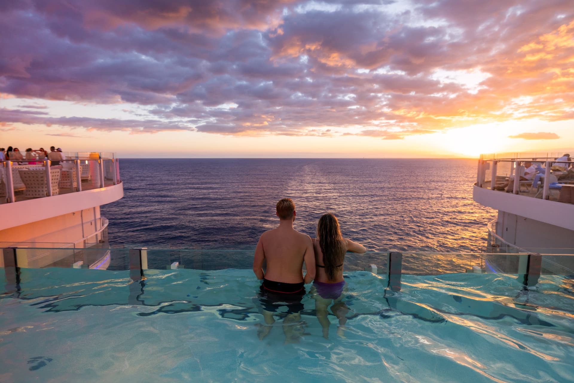 A couple in a ship's infinity pool looking out at the ocean during a colorful sunset