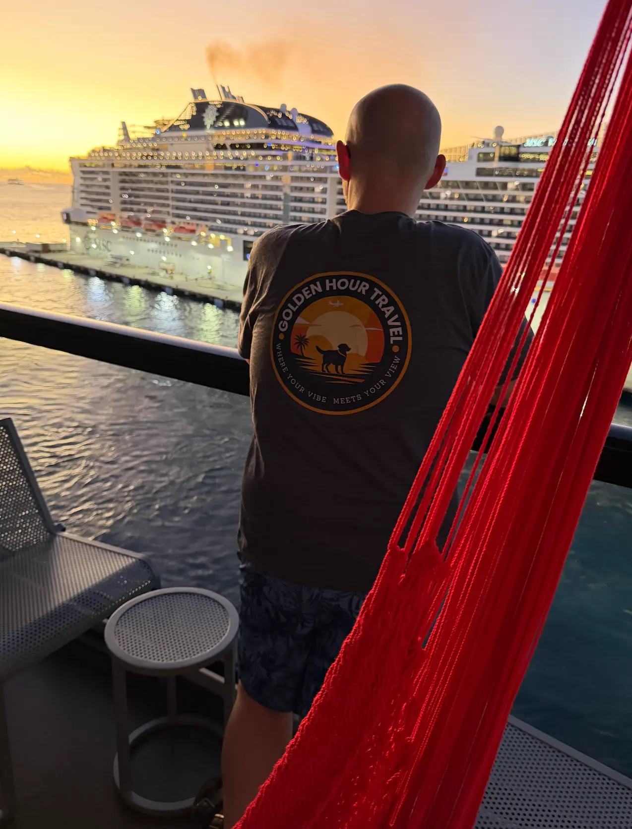 Chris in a Golden Hour Travel shirt on a Virgin Voyages Sea Terrace at sunset, beside a red hammock, looking out at a docked cruise ship