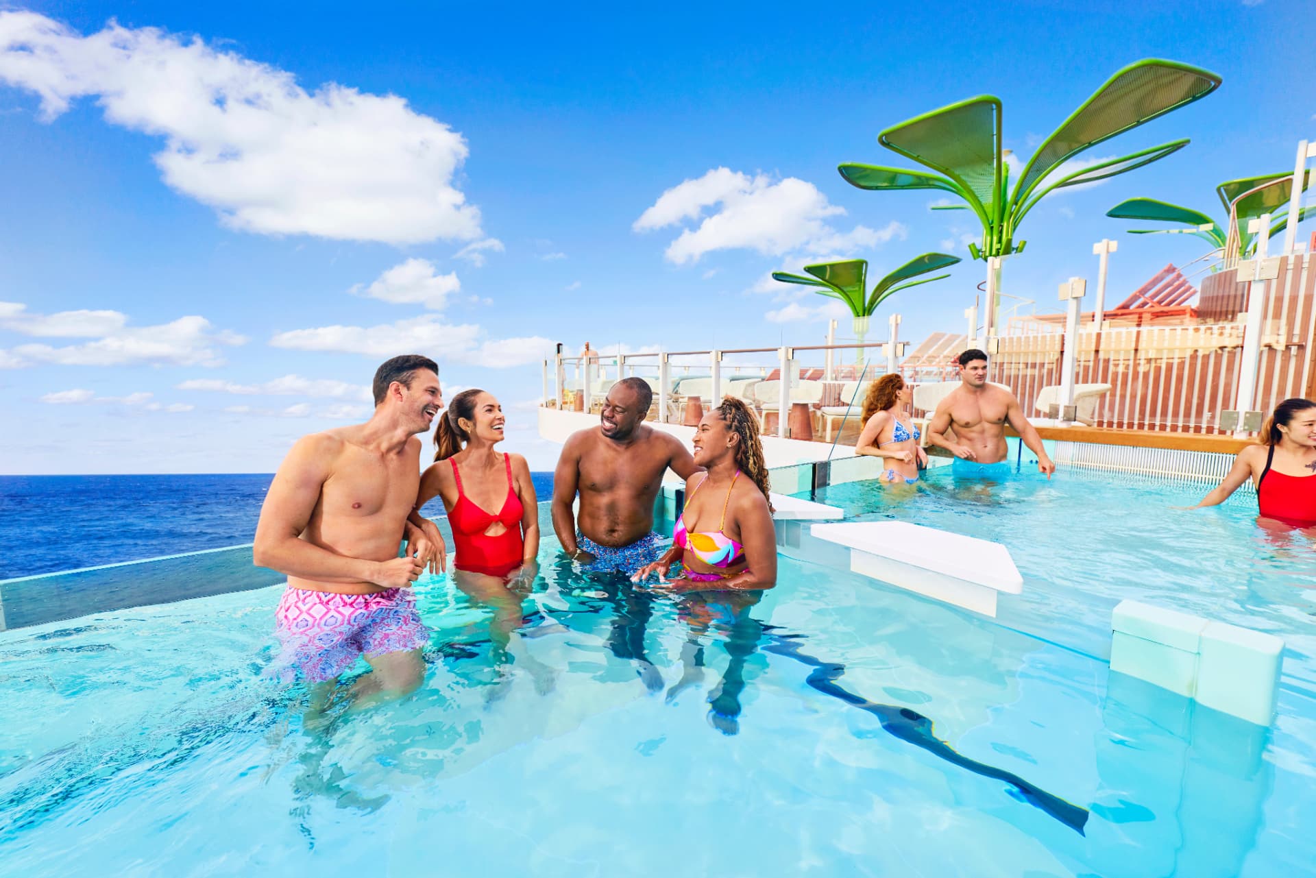 Guests relaxing and talking in a sunlit poolside area on a cruise ship deck overlooking the ocean.