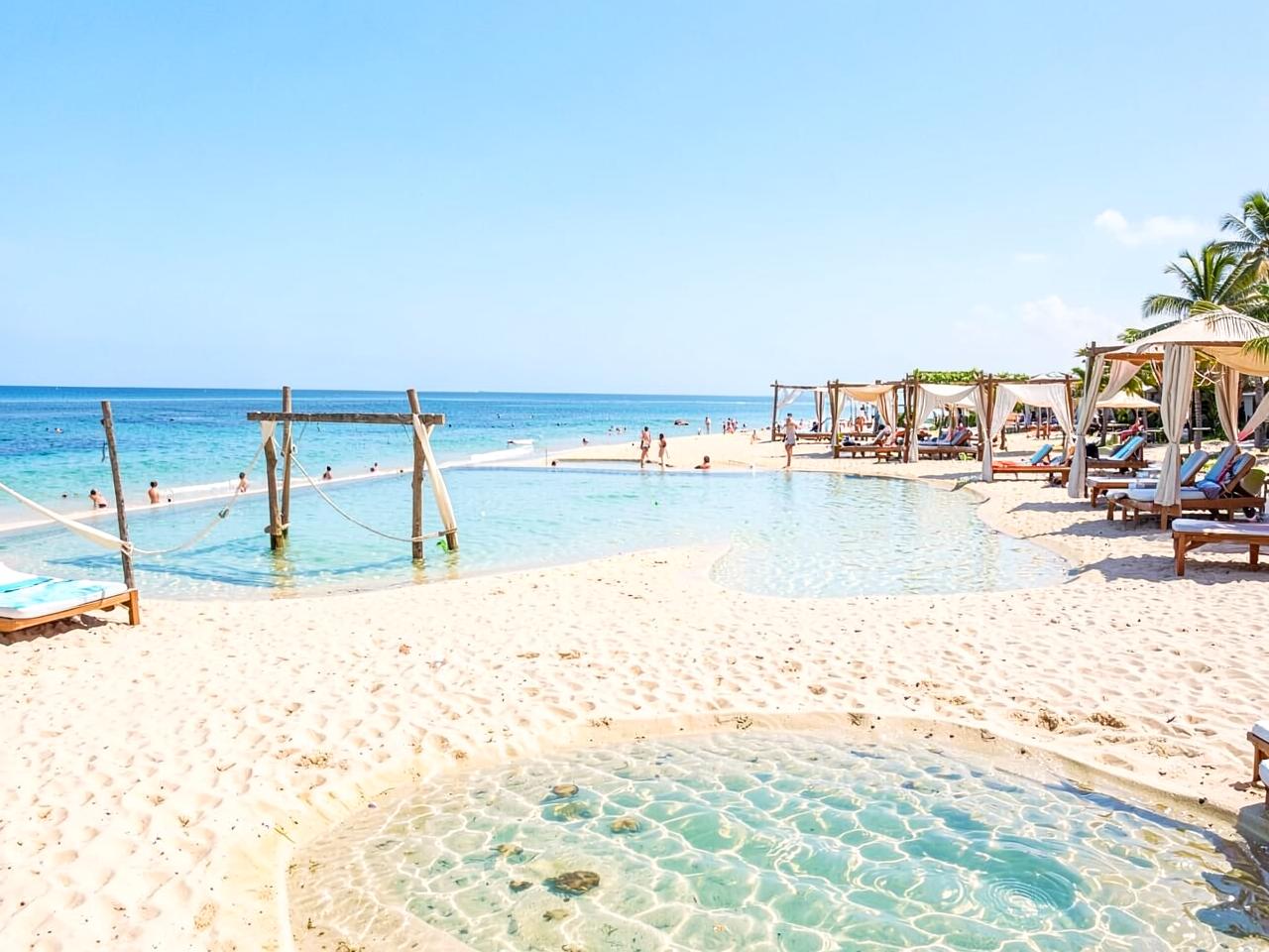 A wide sandy beach with shaded cabanas and a shallow swimming area.